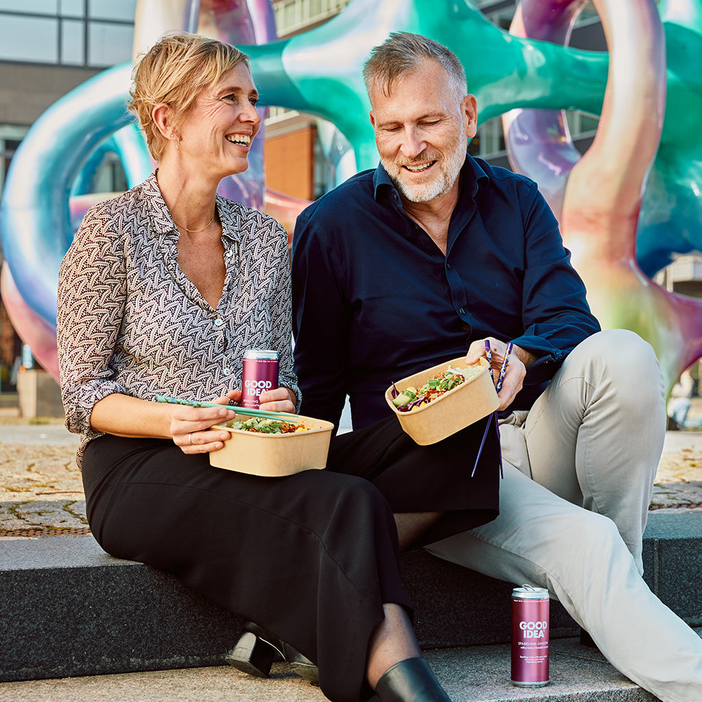 Kvinna och man sitter på en stentrappa och äter mat samt dricker Good Idea sparkling Lingon