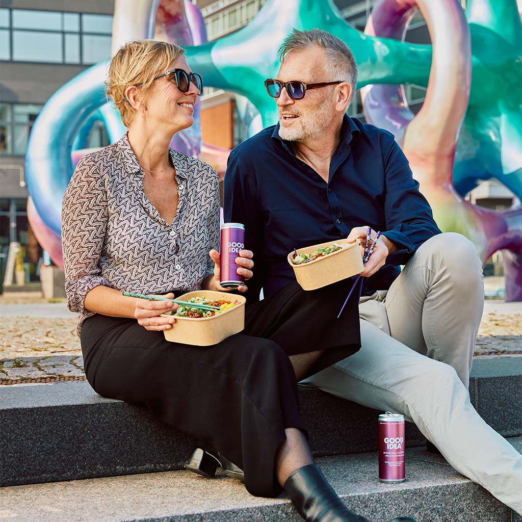 Par sitter på en trappa och äter mat och dricker Good Idea Sparkling Lingon bubbelvatten