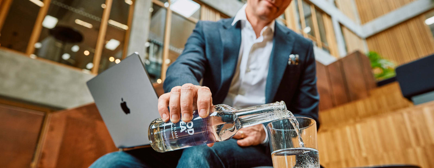 Man i kavaj häller upp en Good Idea sparkling blåbär i ett glas med datorn i sitt knä